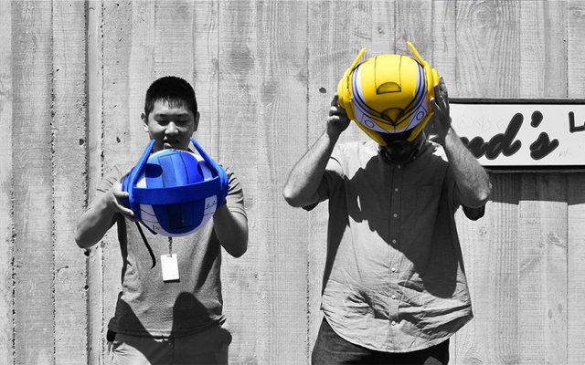A selectively colorized photograph of two men holding robotic helmets. The image is mostly black and white, except for the helmets—one is blue, and the other is yellow—standing out vividly.