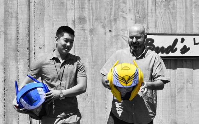 A selectively colorized photograph of two men holding robotic helmets. The image is mostly black and white, except for the helmets—one is blue, and the other is yellow—standing out vividly.